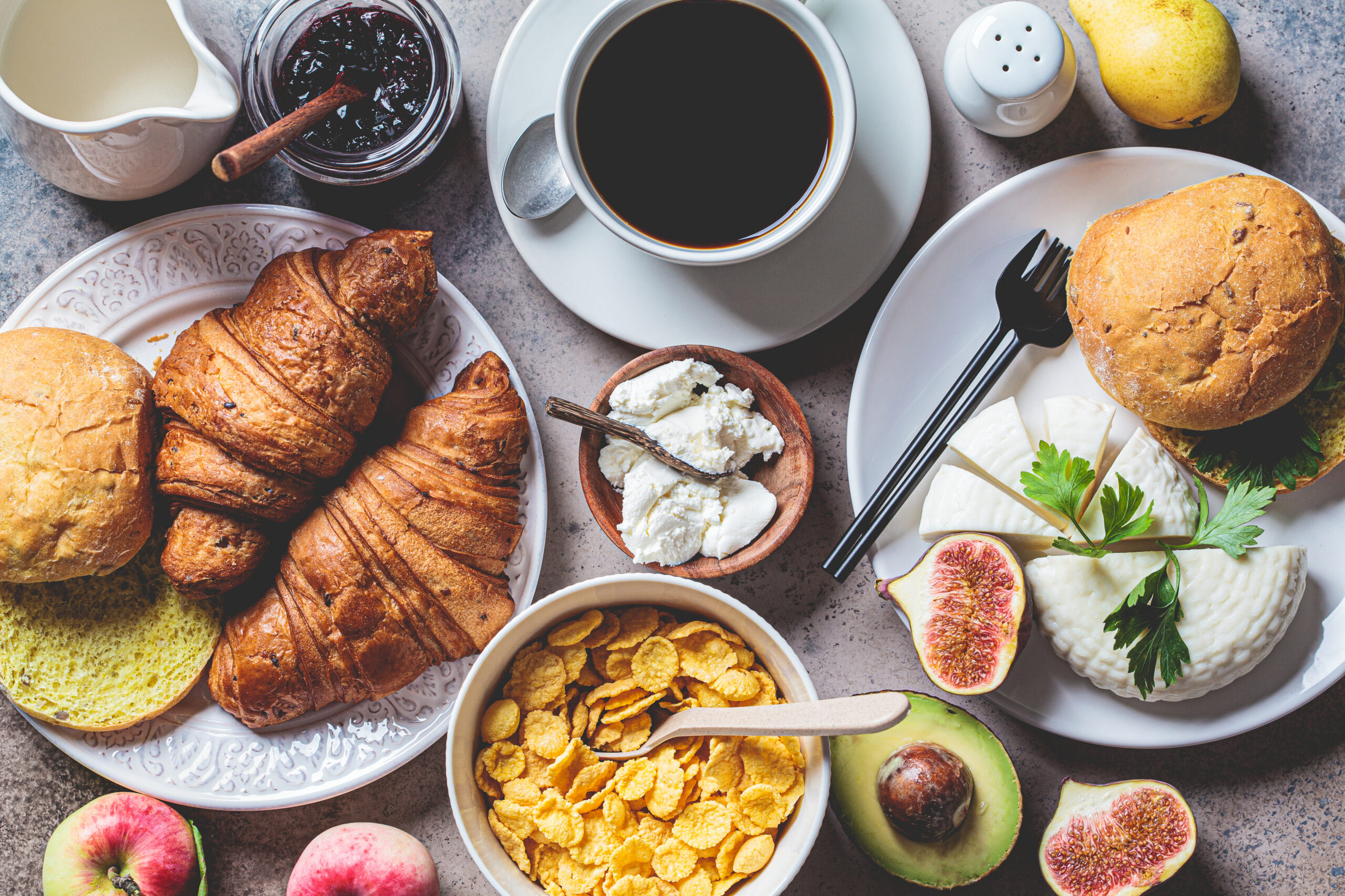 Breakfast table with cornflakes, croissants, fruit, cheese and a cup of coffee. Dark background, top view.